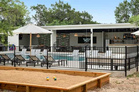 Modern poolside bar and grill with black metal fencing, white umbrellas, and bocce ball court on sandy ground