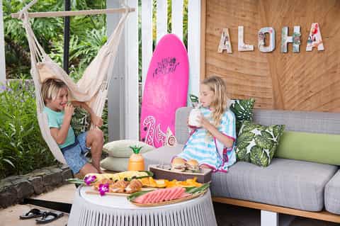 Two children enjoying Hawaiian-themed snacks on a patio with hammock, surfboard, and 'ALOHA' sign