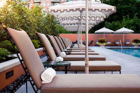 Luxury poolside lounge area with cushioned recliners, towels, striped umbrellas, and city views