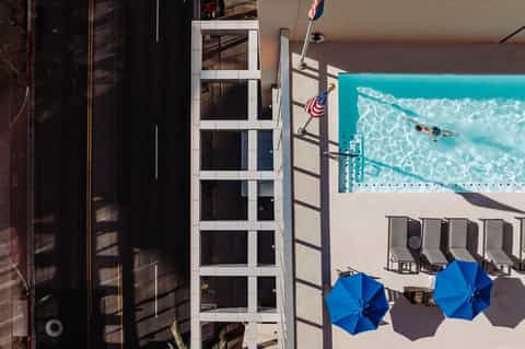 Aerial view of hotel pool with turquoise water, blue umbrellas, lounge chairs, and adjacent building