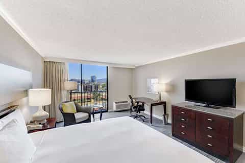 Modern hotel room with king bed, work desk, TV, and balcony overlooking city