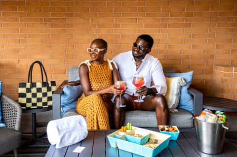 Couple sitting on a brick patio enjoying cocktails with snacks and a beach bag nearby