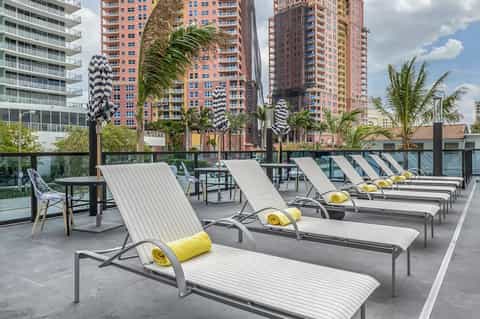 Rooftop lounge with white loungers, yellow pillows, palm trees, and city skyline view