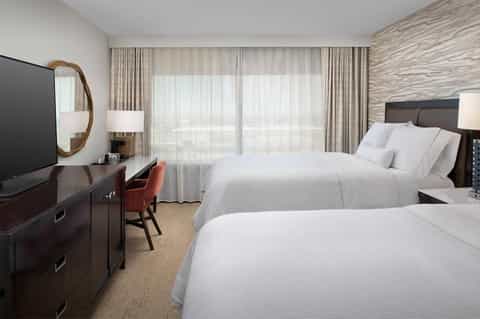 Modern hotel room with two queen beds, dark wood furniture, and floor-to-ceiling curtains