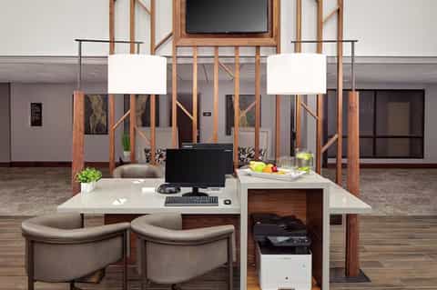 Modern hotel workspace with wooden partitions, pendant lighting, two tan chairs, and a desk with computer monitor