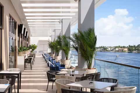 Waterfront dining terrace with white pergola, palm plants, and water views at upscale resort