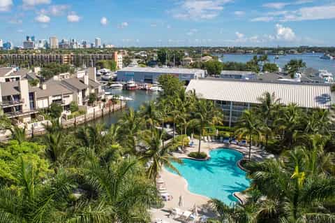 Aerial view of waterfront resort with pool, palm trees, and city skyline