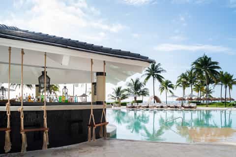 Modern pool bar with rope swing seating, white shade structure, and tropical palm trees by lagoon