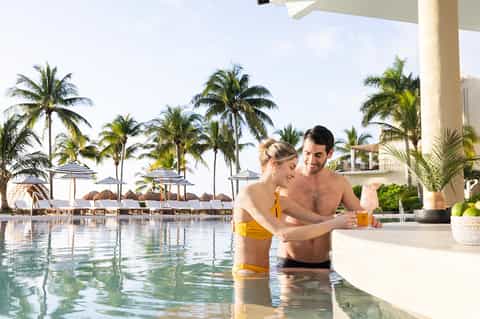 Couple enjoying drinks in a tropical resort pool with palm trees and beach cabanas in background