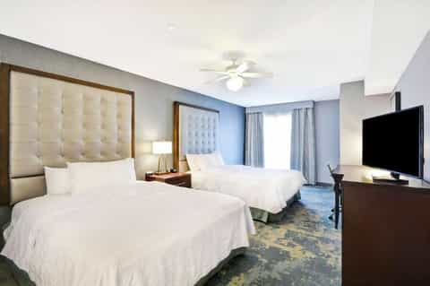 Hotel guest room with two queen beds, cream headboards, flat-screen TV, and blue accent wall