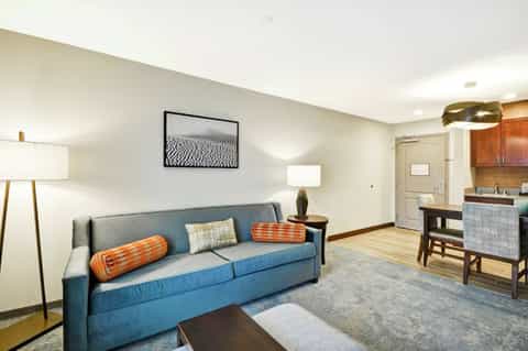 Hotel suite living room with blue sofa, orange pillows, wooden floors, kitchenette, and modern lighting