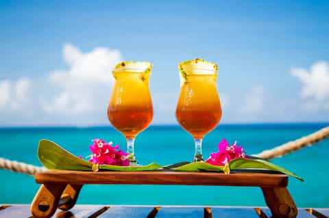 Two tropical cocktails with pineapple garnish on wooden railing overlooking turquoise ocean
