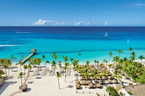 Tropical beach resort with thatched-roof cabana, palm trees, turquoise ocean, and sailboats