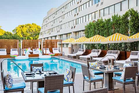 Resort pool deck with yellow-striped umbrellas, lounge chairs, dining tables, and modern white building backdrop