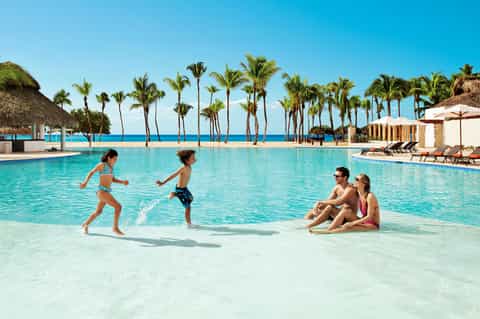 Family playing in shallow turquoise pool with white sand beach and palm trees along horizon