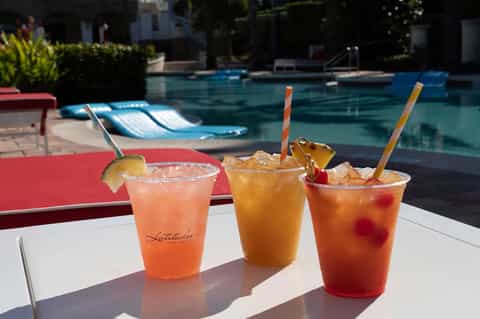 Three colorful beverages with straws on a white table overlooking a resort pool