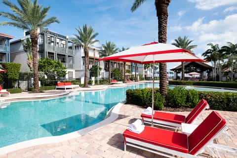 Outdoor resort pool with red and white umbrellas, loungers, and palm trees surrounding modern residential buildings