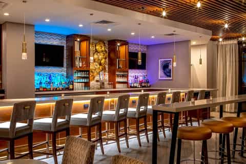 Modern hotel bar with purple accent lighting, wooden bar seating, pendant lights, and decorative display shelves
