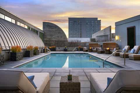 Rooftop pool at dusk with lounge chairs, fire pits, and city skyline backdrop