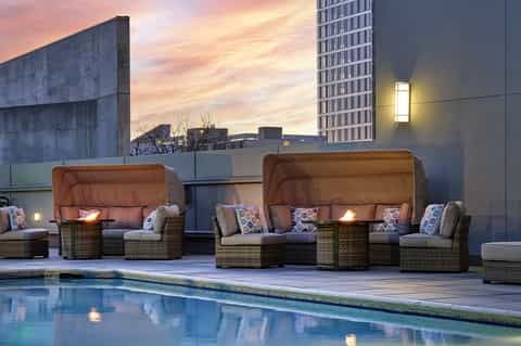 Rooftop pool area at dusk with woven lounge chairs, fire pits, and city skyline