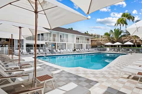 Resort pool area with white umbrellas, lounge chairs, and a two-story beachfront building under blue sky