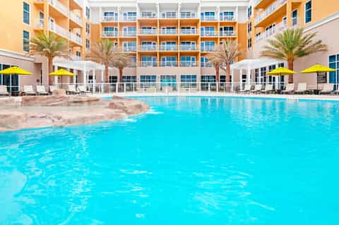 Resort courtyard with turquoise swimming pool, orange and white building, palm trees, and yellow umbrellas