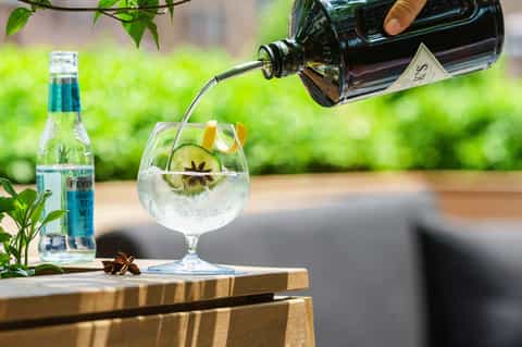 Gin and tonic cocktail being poured into a glass with cucumber and star anise garnish on wooden table