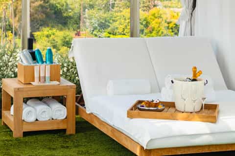 Spa lounge chairs with white cushions beside wooden side table holding rolled towels and amenities