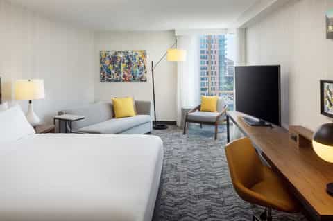 Modern hotel room with white bed, gray seating area, yellow accents, and city view through windows