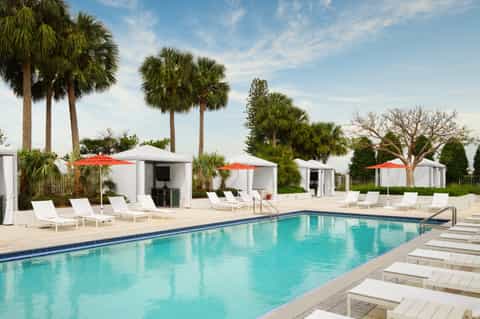 Resort pool with white cabanas, red umbrellas, and palm trees under blue sky