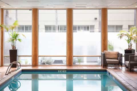 Indoor lap pool with large windows, wooden columns, potted plants, and lounge seating area