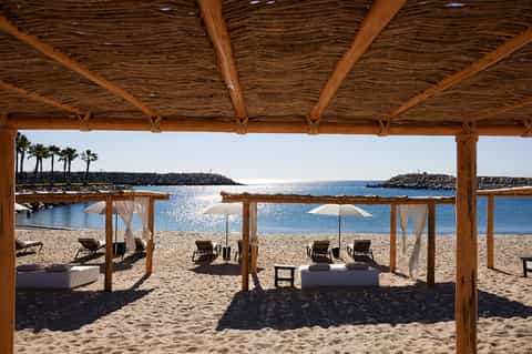 Beach cabanas with wooden pergolas, lounge chairs, and umbrellas facing clear blue ocean water