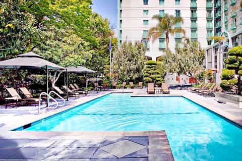 Resort pool with clear turquoise water, black umbrellas, lounge chairs, palm trees, and white high-rise building