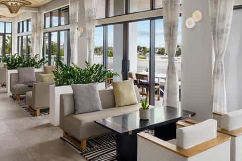 Bright, modern lounge with beige sofas, large windows overlooking city, indoor plants, and contemporary light fixtures
