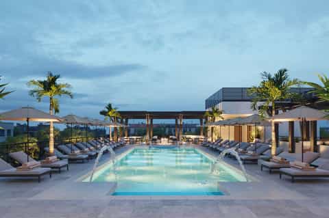 Luxury rooftop pool at dusk with loungers, palm trees, modern architecture, and city skyline views