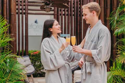 Couple in spa robes toasting with champagne glasses in a tropical courtyard setting