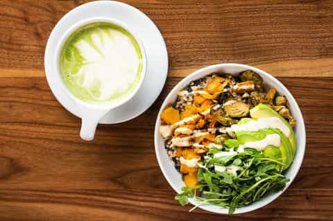 Healthy bowl with avocado, roasted vegetables, and greens beside matcha latte on wood table