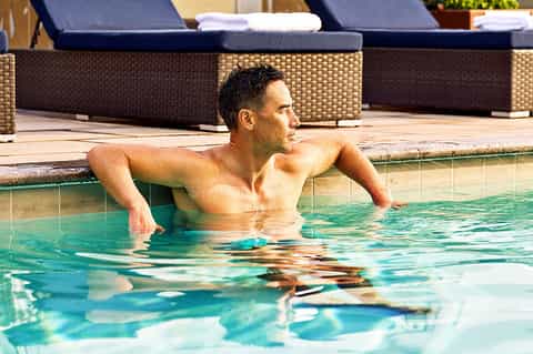 Man relaxing in resort pool with lounge chairs and dark cushioned seating in background