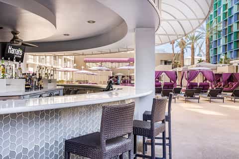 Modern hotel bar with curved white counter, rattan chairs, and purple cabana seating area overlooking palm trees