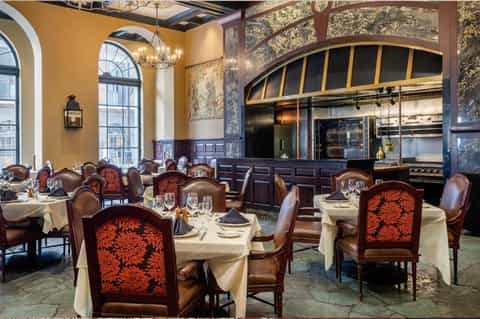 Upscale fine dining restaurant with gold walls, arched windows, open kitchen, and tables set with cream linens and red patterned chairs
