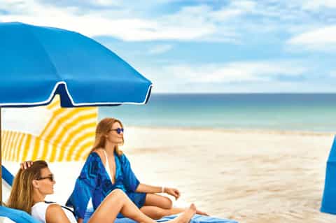 Two women relaxing under blue and yellow beach umbrellas with ocean view in background