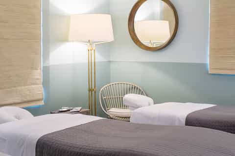 Modern hotel bedroom with two beds, soft lighting, wooden wicker chair, and light blue accent wall