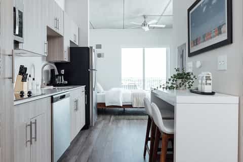 Compact studio hotel room with kitchenette, white bedding, desk area, and floor-to-ceiling windows.