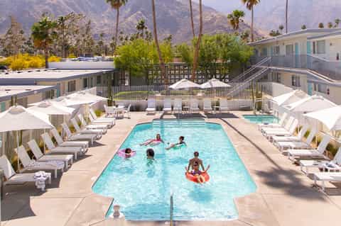 Resort pool with families swimming, palm trees, mountains in background, white umbrellas and loungers