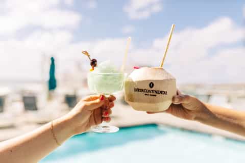 Two tropical cocktails with straws held at a rooftop pool overlooking a coastal city