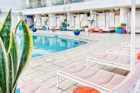 Resort pool deck with pink lounge chairs, colorful planters, white cabanas, and palm trees