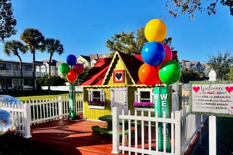 Bright yellow playhouse with red roof, white fence, and large colorful balloons in residential community setting