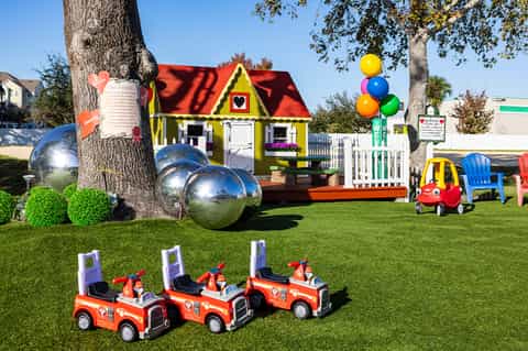 Colorful children's play area with yellow house, red fire trucks, and metallic spheres on lawn
