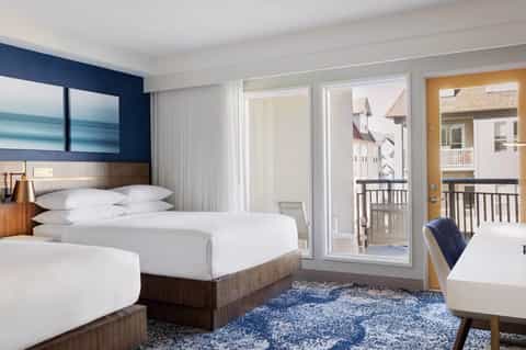 Modern hotel room with two beds, dark blue accent wall, ocean artwork, and balcony overlooking coastal homes