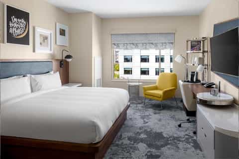 Modern hotel bedroom with queen bed, yellow armchair, desk area, and city views through large window